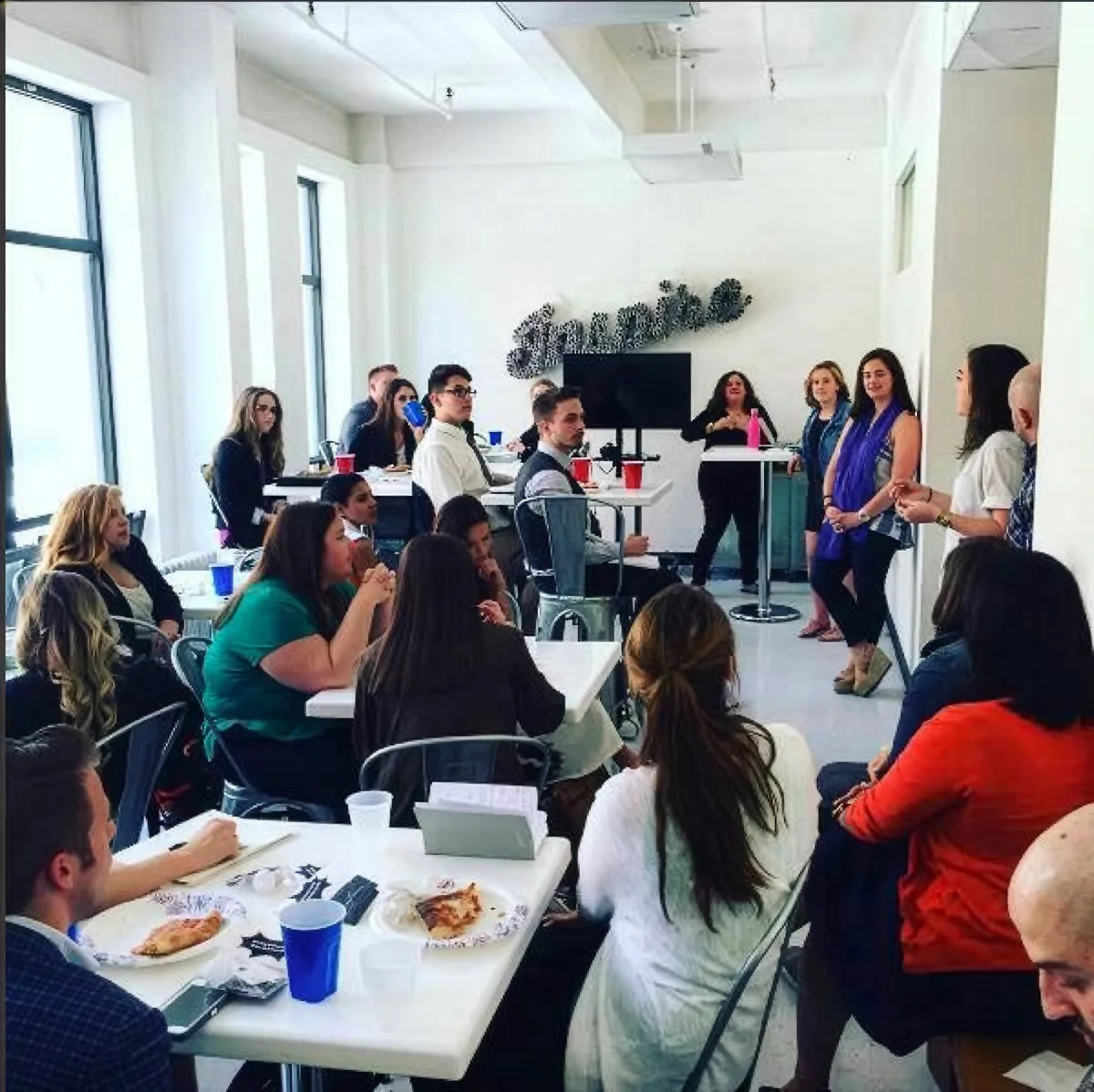People seated at white tables with pizza and drinks in a bright office, others standing near a screen and a cursive wall sign.
